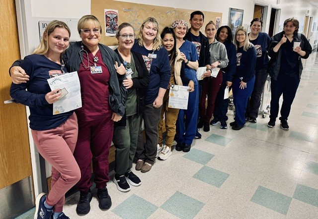 Group of medical staff and students in scrubs lined up in a hallway, several holding certificates and smiling at the camera.