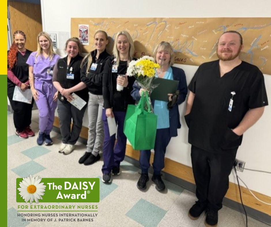 Group of nurses pose in a hallway with The DAISY Award logo, celebrating an award; one holds a bouquet and a green gift bag.