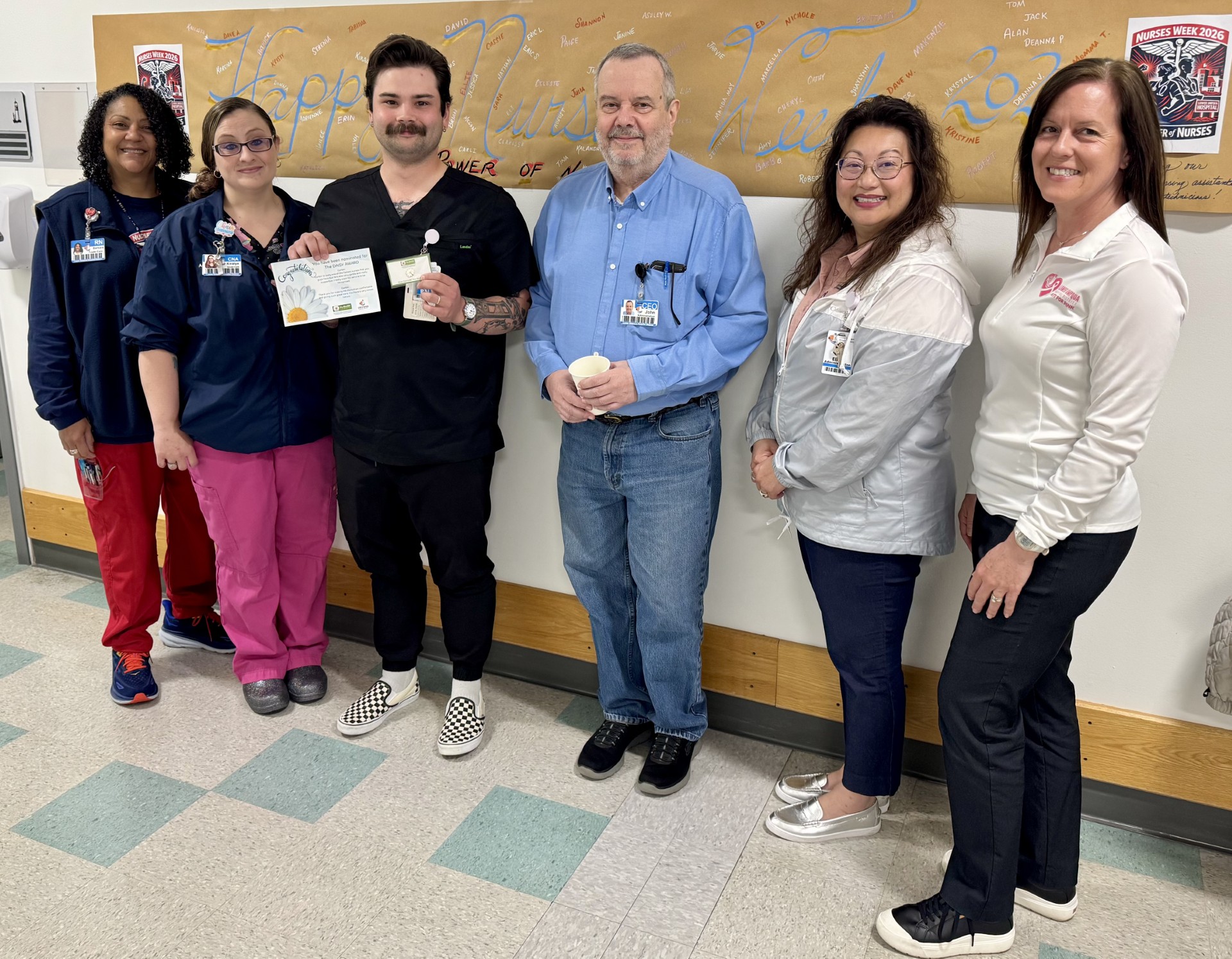 Six hospital staff posing in a hallway with a signed banner behind them; one person in the center holds a certificate and badge, others wear scrubs or uniforms and smile at the camera.