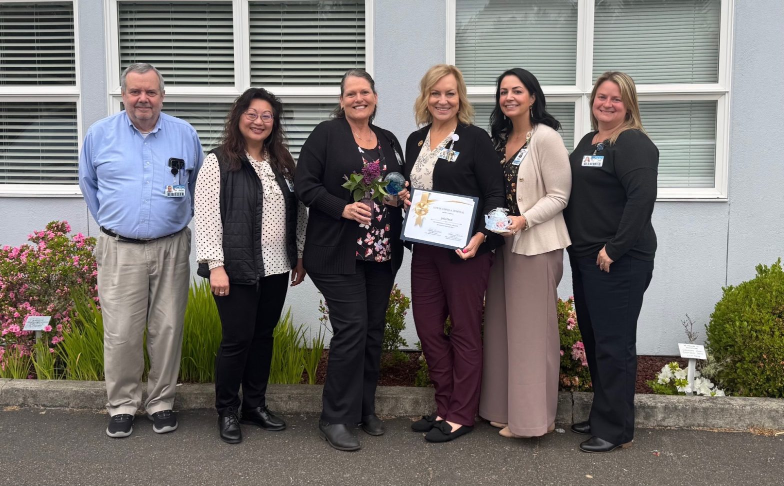 Six professionals pose outdoors in front of a building, one holds a framed certificate and another a glass award with flowers nearby in bloom.