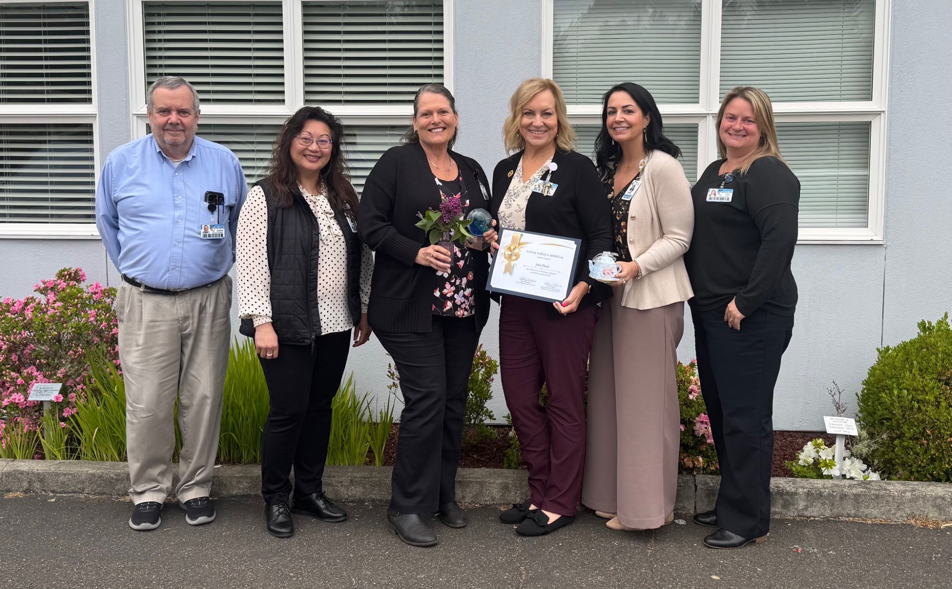 Six professionals pose outdoors in front of a building, one holds a framed certificate and another a glass award with flowers nearby in bloom.