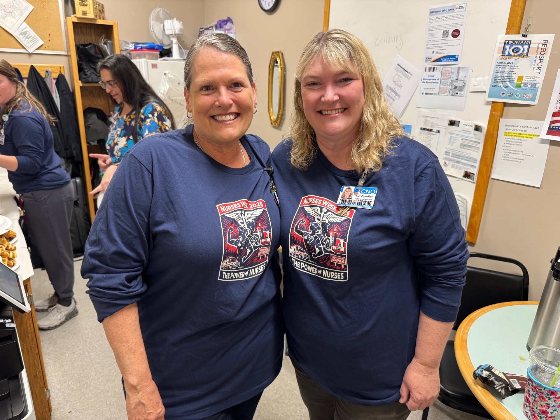 Two smiling nurses in matching navy shirts with the Nurses Week logo, posing in a staff break room.