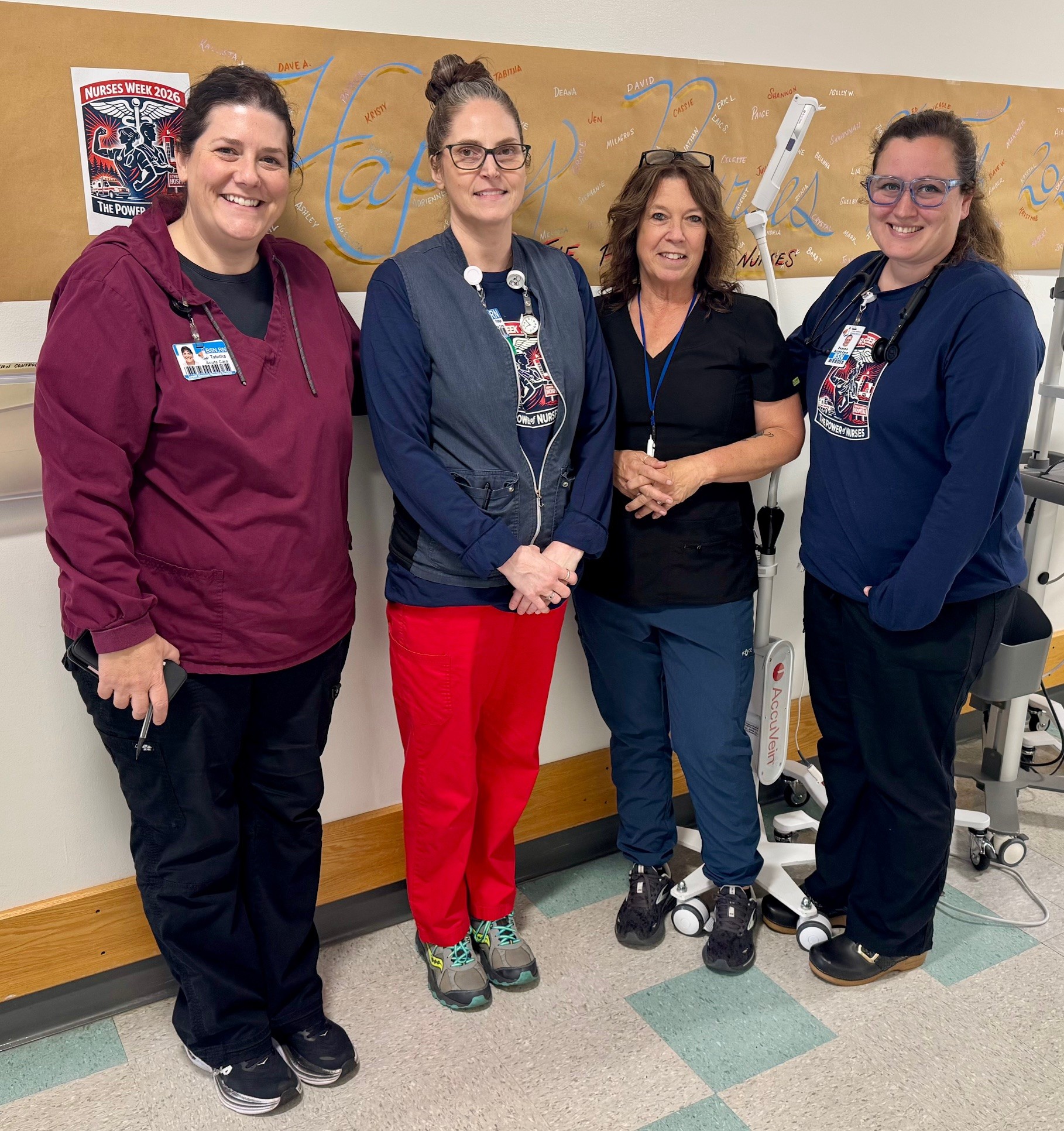 Four nurses posing together in a hospital hallway with a Nurses Week poster on the wall. Four women in scrubs smile at the camera, medical equipment nearby.