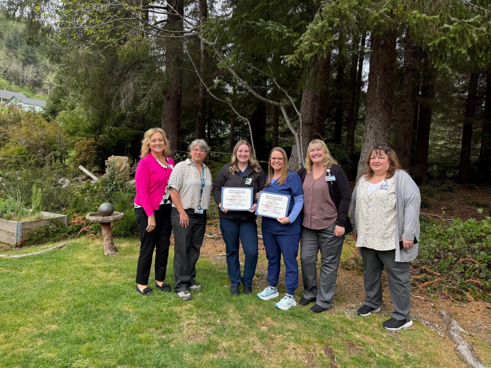 Six women stand together in a grassy yard with tall trees behind them; two in the center hold framed certificates, smiling at the camera.