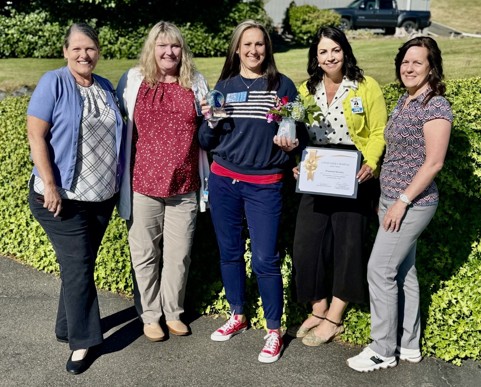 Group of six women standing together outdoors, smiling, with one holding a trophy and another a framed certificate.