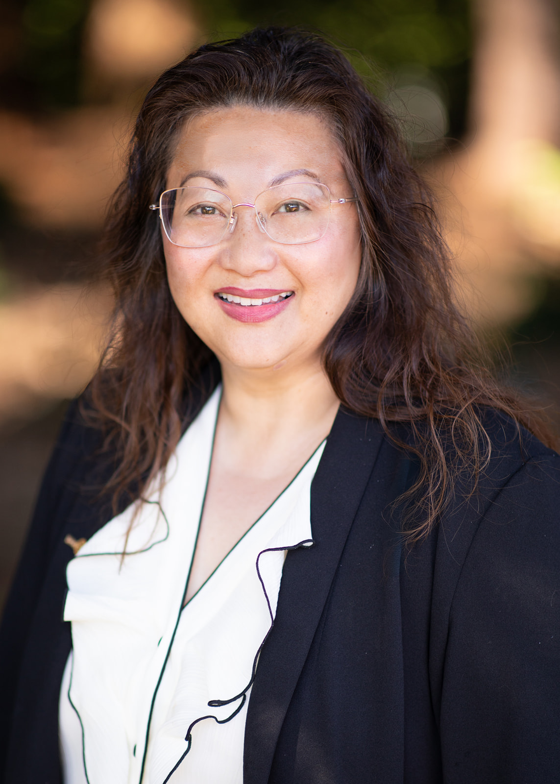 Smiling woman with round glasses and wavy hair wearing a white blouse and black blazer, outdoors for a professional headshot.