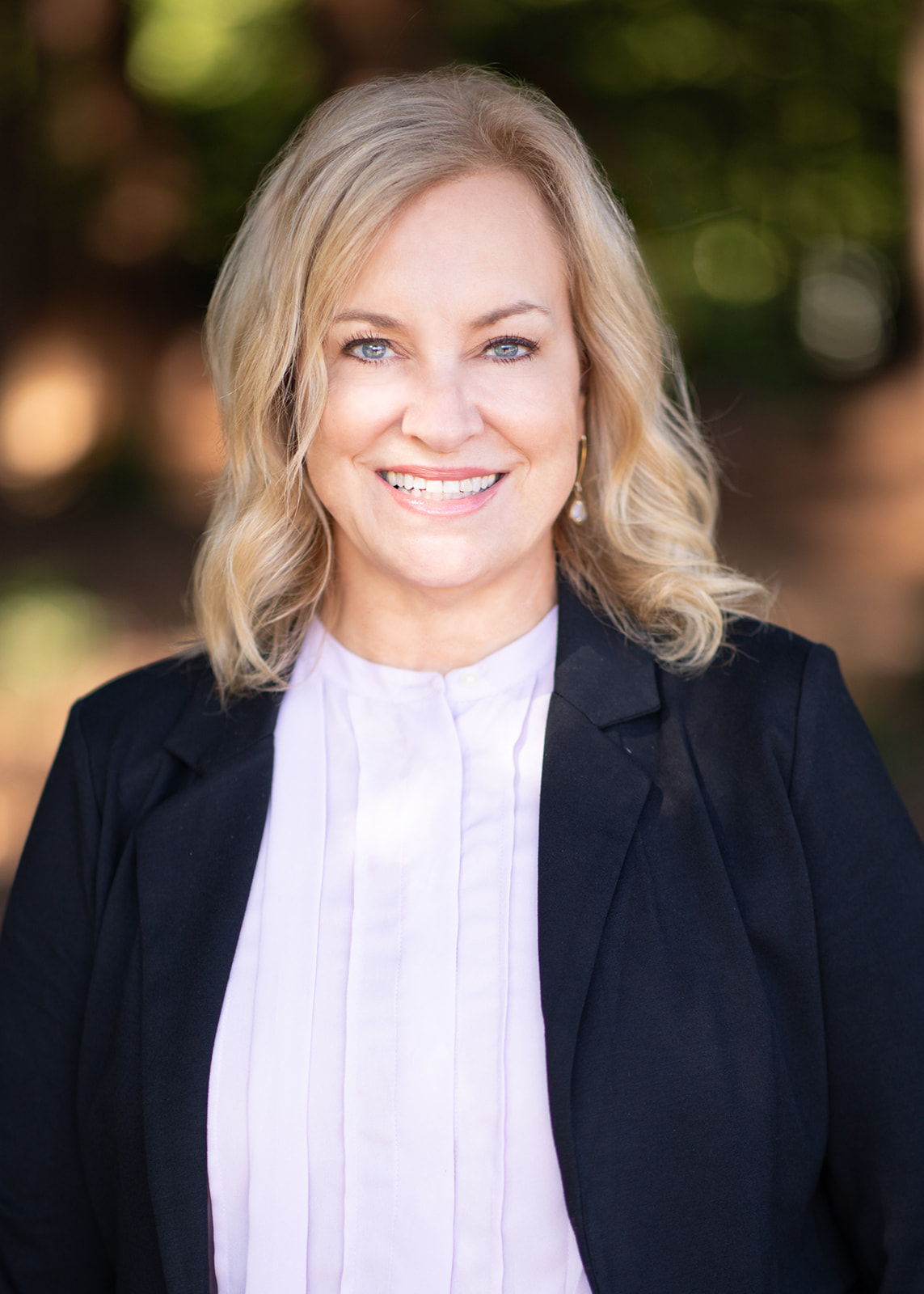 Professional headshot of a smiling blonde woman in a black blazer and lavender blouse, outdoors with greenery in the background.
