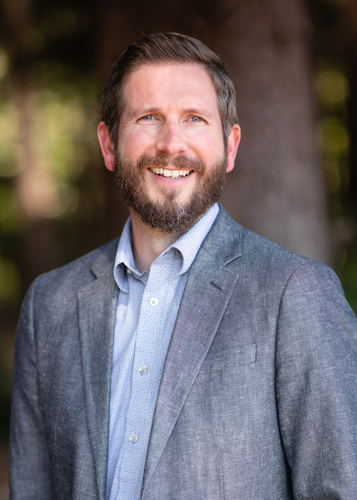 Smiling man with a beard in a gray blazer and light blue shirt, outdoors with a blurred park background.