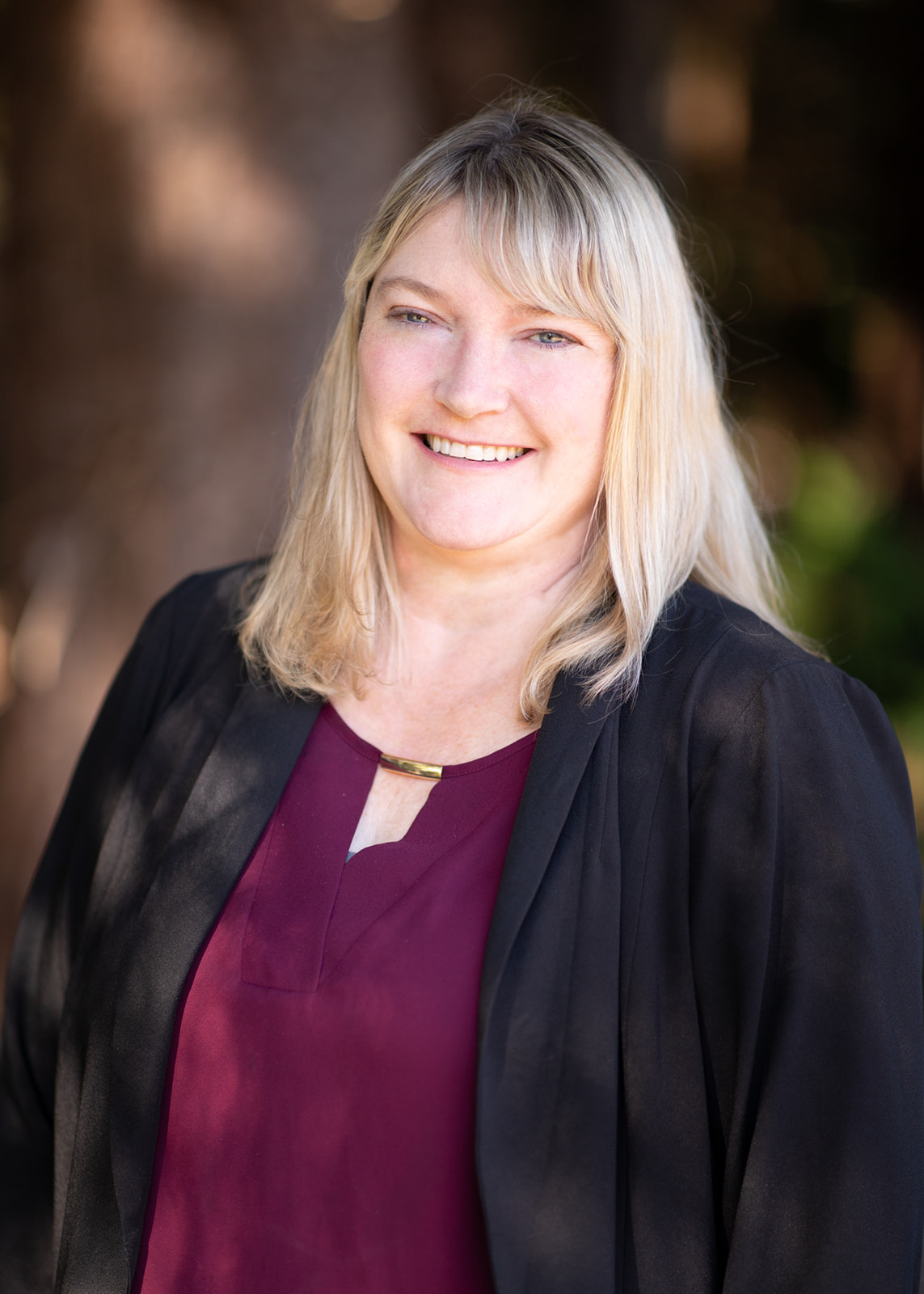 Professional headshot of a smiling blonde woman wearing a burgundy blouse and black blazer outdoors, with a blurred natural background.