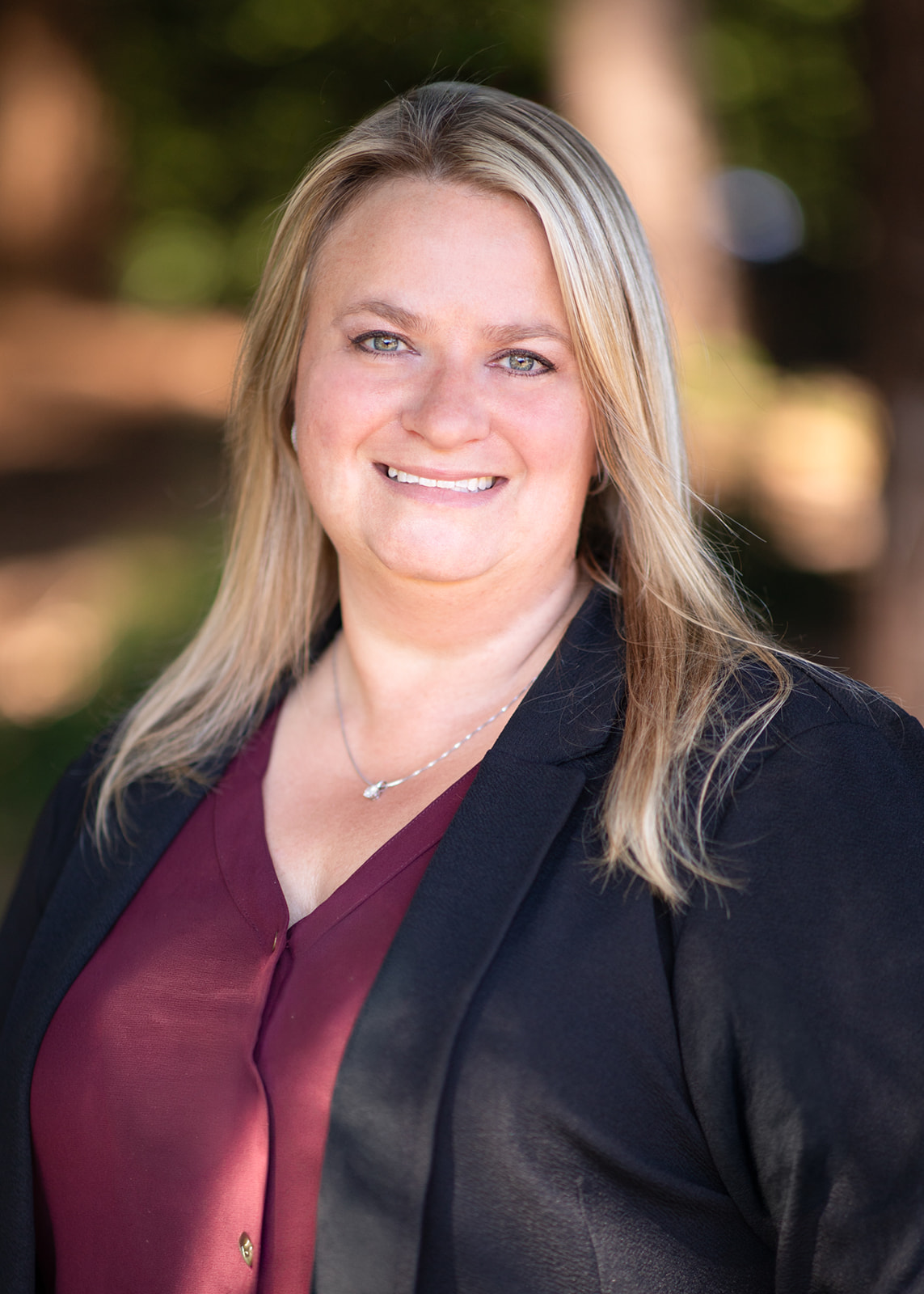 Professional headshot of a smiling woman with blonde hair, wearing a burgundy blouse and black blazer outdoors.