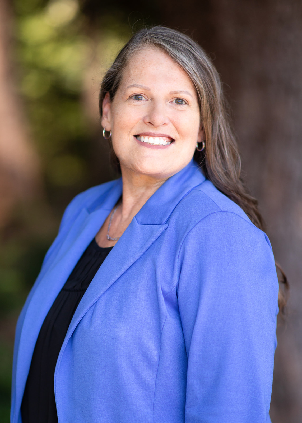 Smiling woman in a blue blazer outdoors, head-and-shoulders portrait on a blurred background.