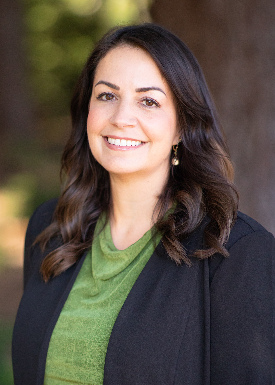 Professional headshot of a smiling woman with dark hair, wearing a green blouse and black blazer, outdoors.