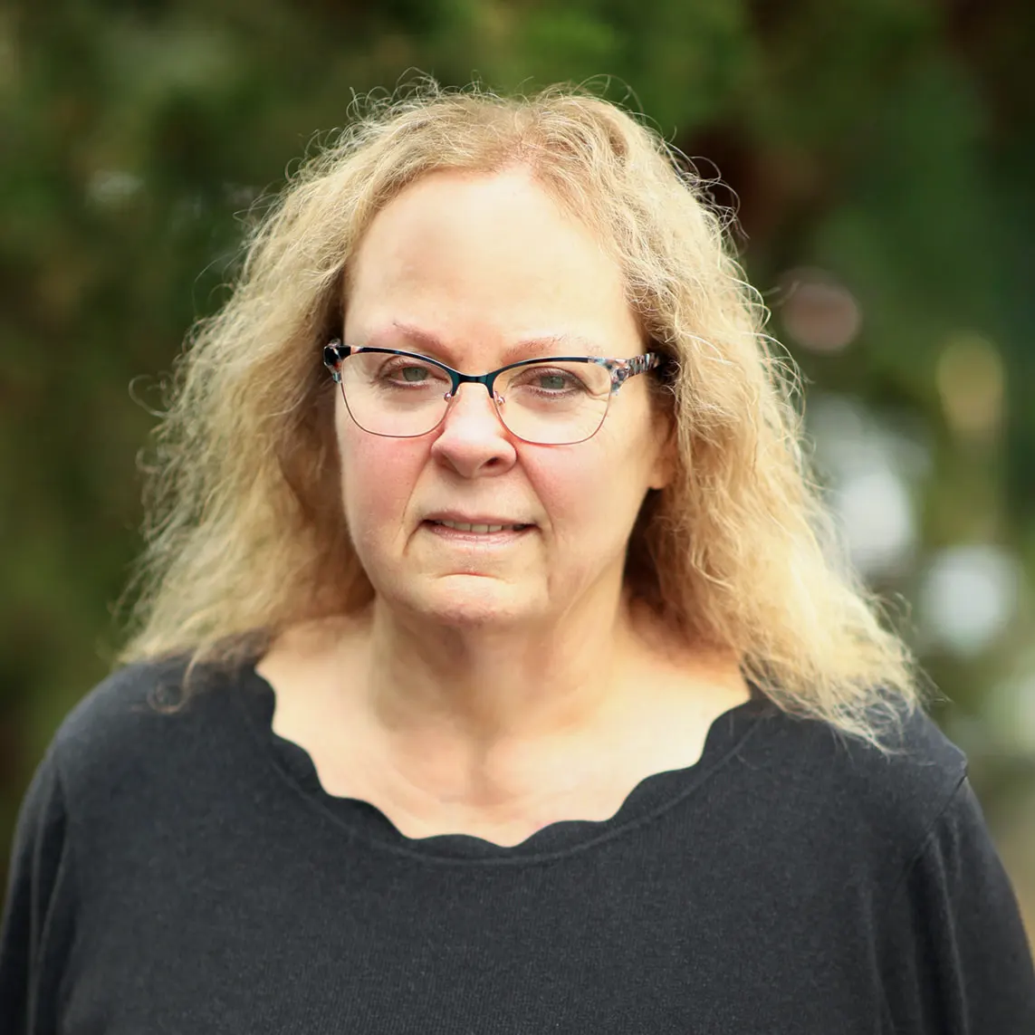 Brenda Fraley_websize Portrait of a middle-aged woman with wavy blonde hair and glasses, standing outdoors in a black top.