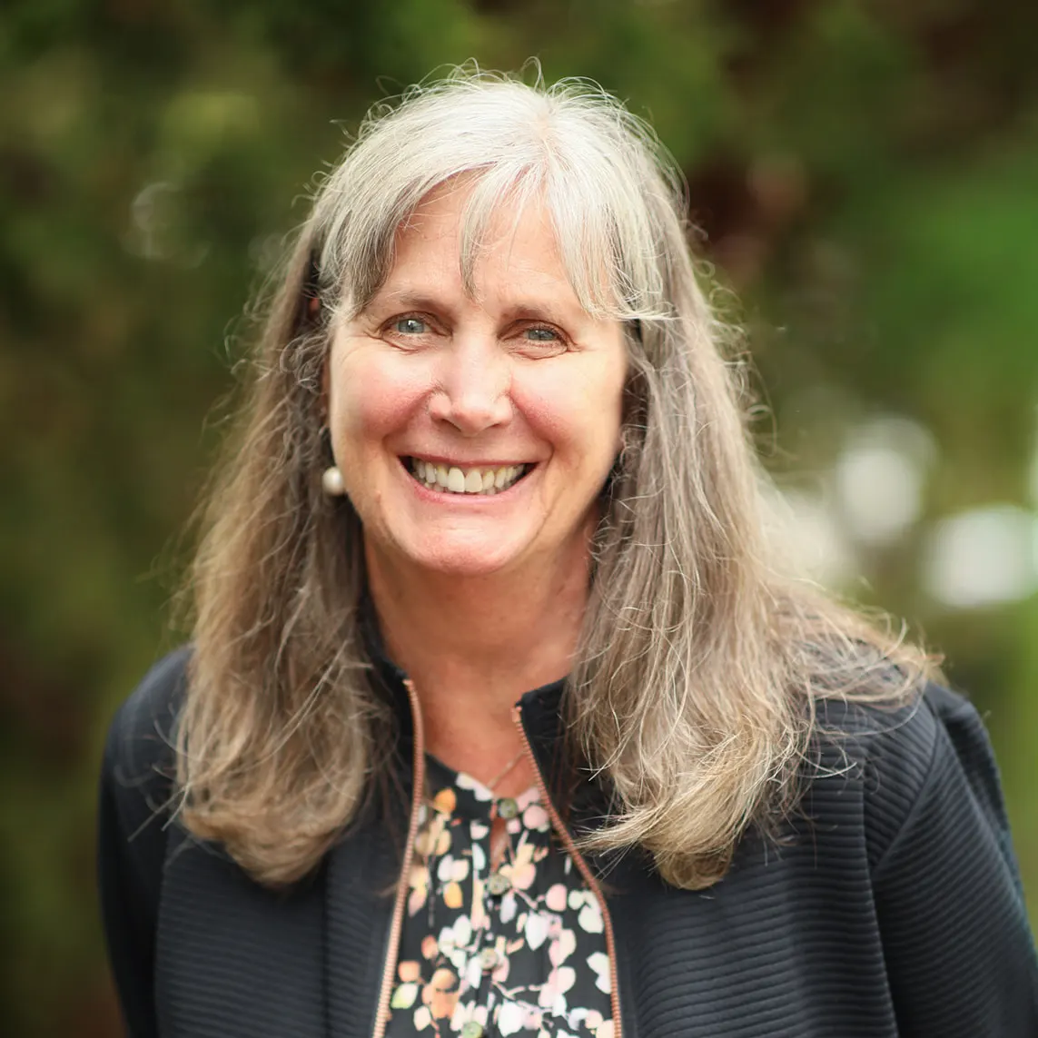 Cheryl Young_websize Smiling woman with long gray hair outdoors, wearing a black jacket and floral blouse.
