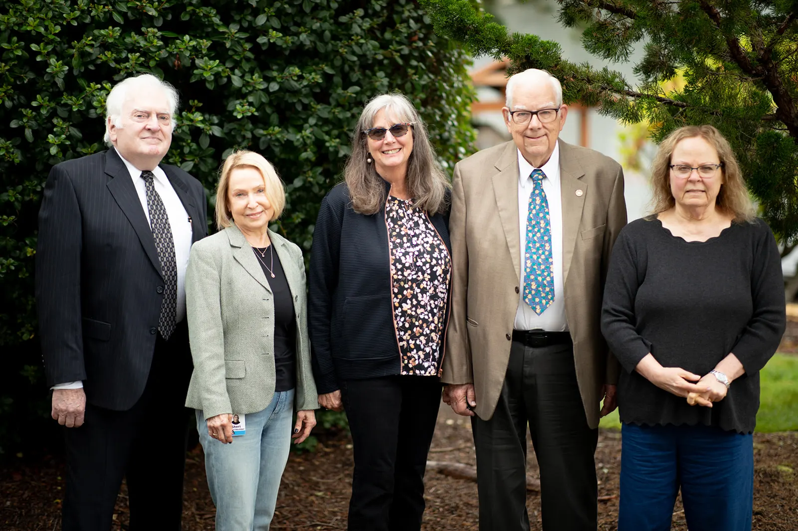 Five adults stand together outdoors in front of shrubs, dressed in business and smart casual attire.