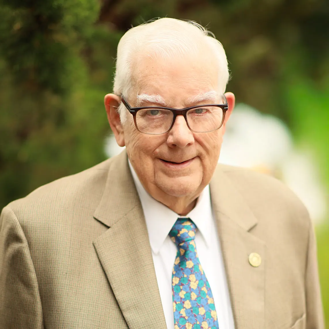 Ron Kreskey_websize Portrait of an elderly man in a tan suit and colorful floral tie, outdoors with greenery in the background.