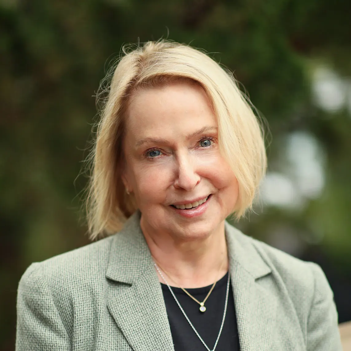 Sheri Aasen_websize Portrait of a smiling older woman with short blonde hair, wearing a gray blazer and black top outdoors.